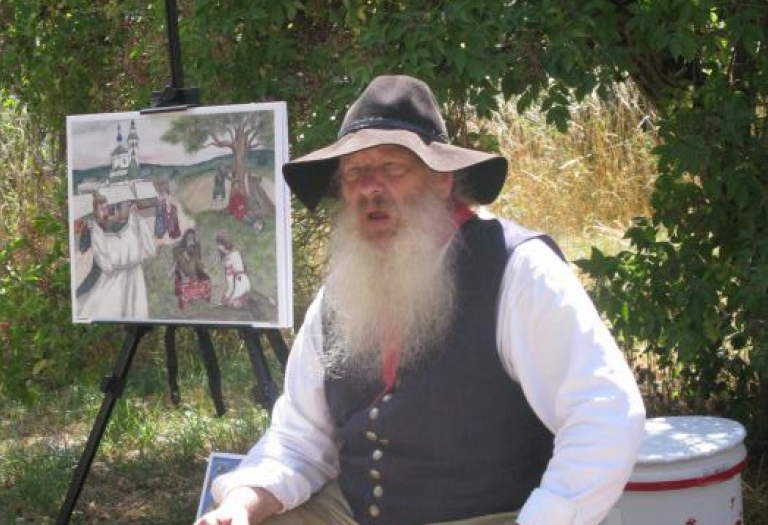 Ein Mann mit langem weißen Bart und großem Filzhut sitzt im Freien vor einem mittelalterlichen Bild.